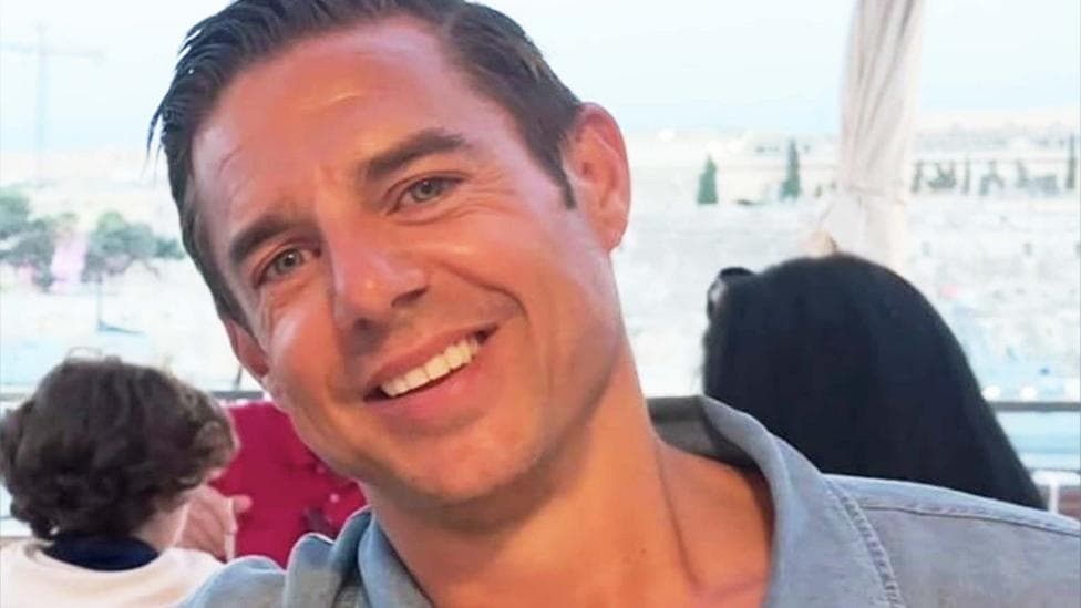 Close-up headshot of a smiling Nicholas Brandram with short dark hair, blue eyes. He is clean-shaven. He is wearing a blue open-collared shirt outdoors, with a blurred background showing an outdoor seating area and buildings under a clear sky.