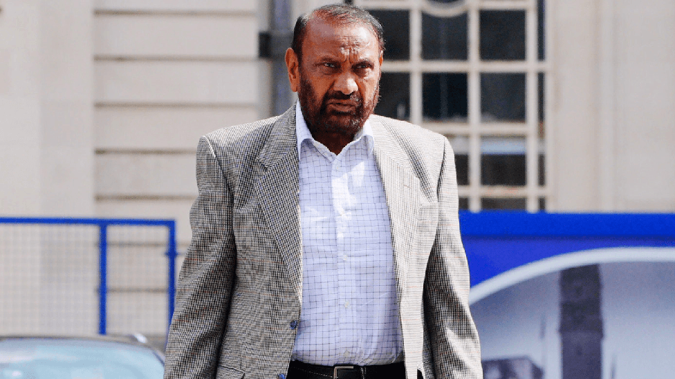 A man with a grey jacket and white shirt walking away from a building.