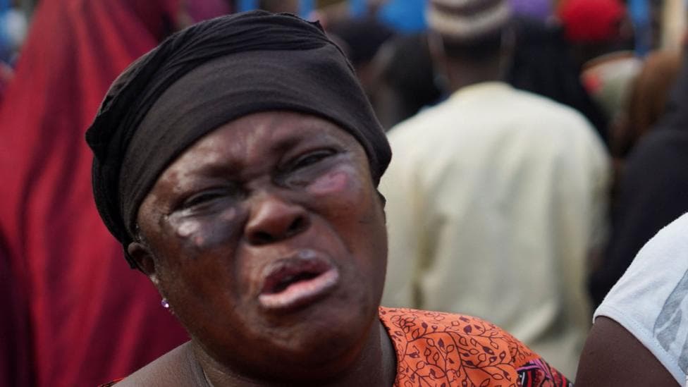 A close-up photo of a woman crying. She has a black covering on her hair and an orange top.