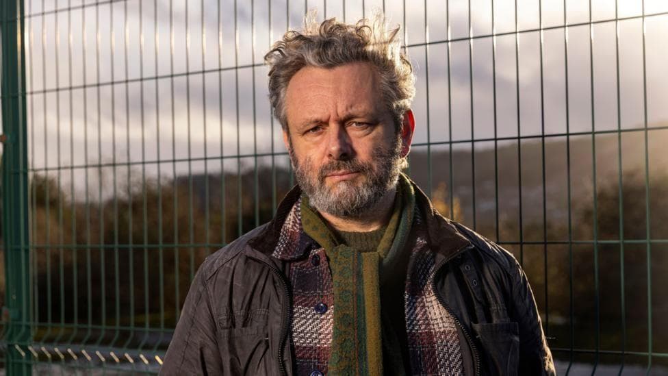 Michael Sheen looks at the camera. He is wearing a green scarf and a plaid red shirt and a coat over the top. Behind him is a waste site which is fenced off.