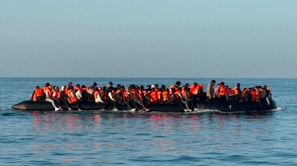 A large black inflatable dinghy carrying dozens of people wearing orange life jackets travels across calm sea water. People are seated along both sides of the vessel and across its centre.