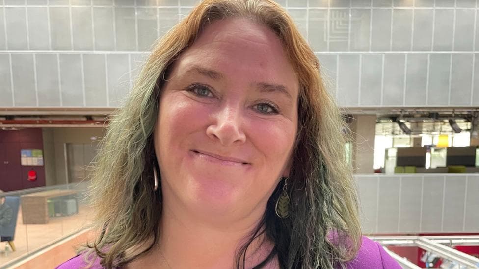 Cara McKee standing indoors in a modern atrium-style building, wearing a purple top, leaf-shaped earrings and a matching pendant necklace, facing the camera with glass railings and an open interior space visible in the background.