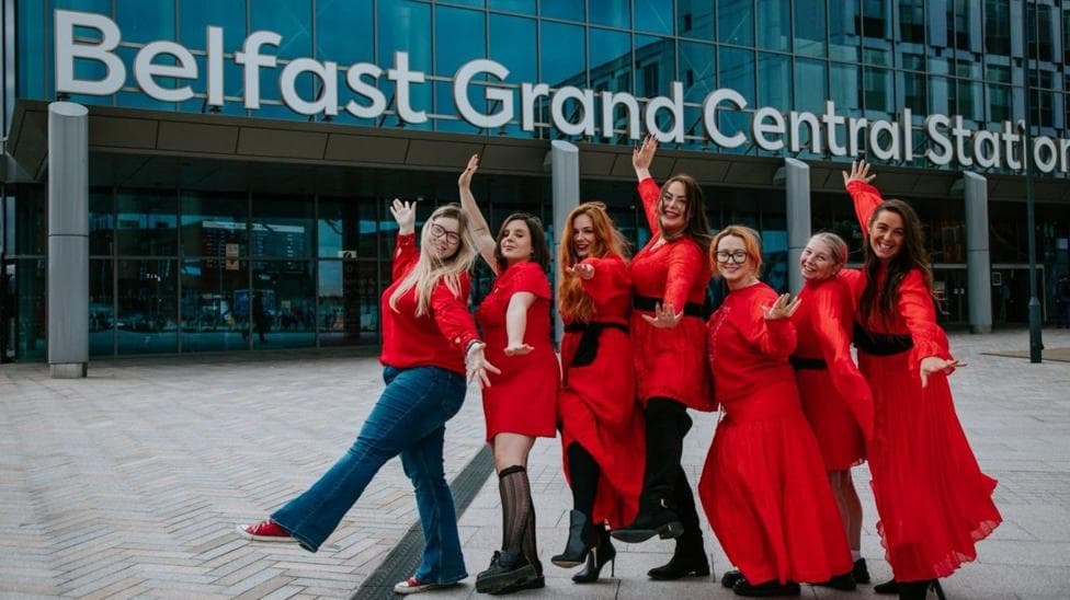 Seven women in red dresses and tops wave in front of Belfast Grand Central Station to promote the Cathy's Call event for Culture Night Belfast.