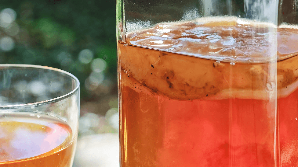 A jar of light tea coloured kombucha is on the right of the photo with a scoby floating on top of it. The the left is a partly-seen glass containing kombucha.