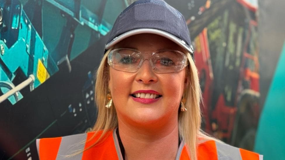 A woman with longish blond hair, gold earrings and a protective glasses and a high vis jacket smiles at the camera.