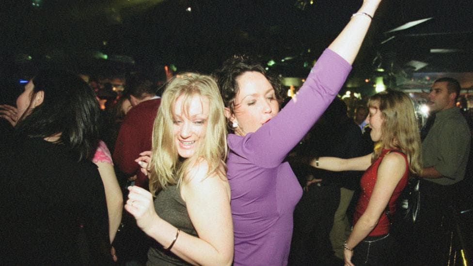 Two women stand back to back while dancing in a nightclub. Other dancers are visible in the background. One of the woman has blond hair and a grey or green top. The other woman has dark hair, is wearing a purple top and is waving one arm in the air.