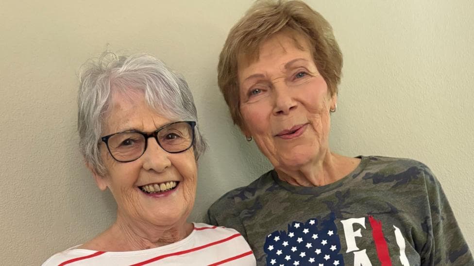 Kathleen Wilson and Shirley Yaeger are standing together and smiling at the camera. Kathleen has short, grey hair and black-rimmed glasses. She is wearing a white top with thin red stripes. Shirley has short, brownish hair and is wearing a camouflage T-shirt featuring aspects of the American flag