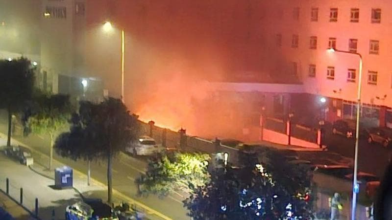 A fire in the basement of a hotel. Flames can be seen from below the building, and emergency services can be seen in the foreground.