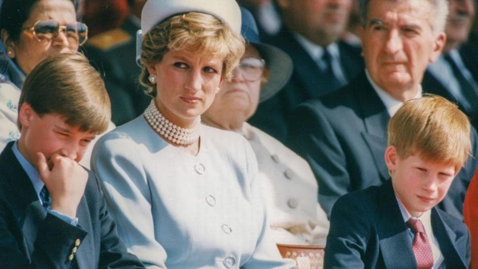 Princess Diana, wearing a light blue dress and hat, sits in between a young Prince William on the left (holding his hand to his mouth) and Harry to the right. Diana is looking into the distance to the left.