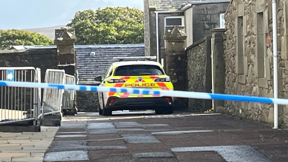 Police tape stretches across the foreground of a narrow lane between stone buildings, with a marked police car parked beyond the cordon and temporary barriers positioned along one side of the passage.