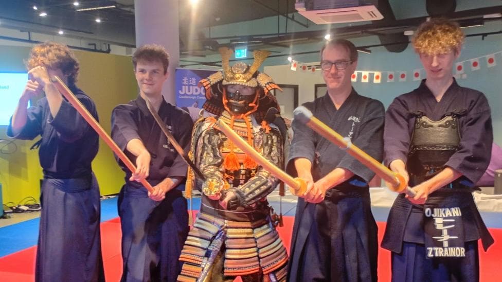 Five people are standing in a row holding Japanese swords. The person in the centre is wearing traditional Japanese multi-coloured armour. The other four are wearing blue clothing