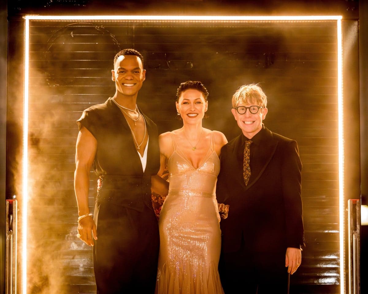 Strictly presenters Johannes Radebe, Emma Willis and Josh Widdicombe smile together in a lit doorway