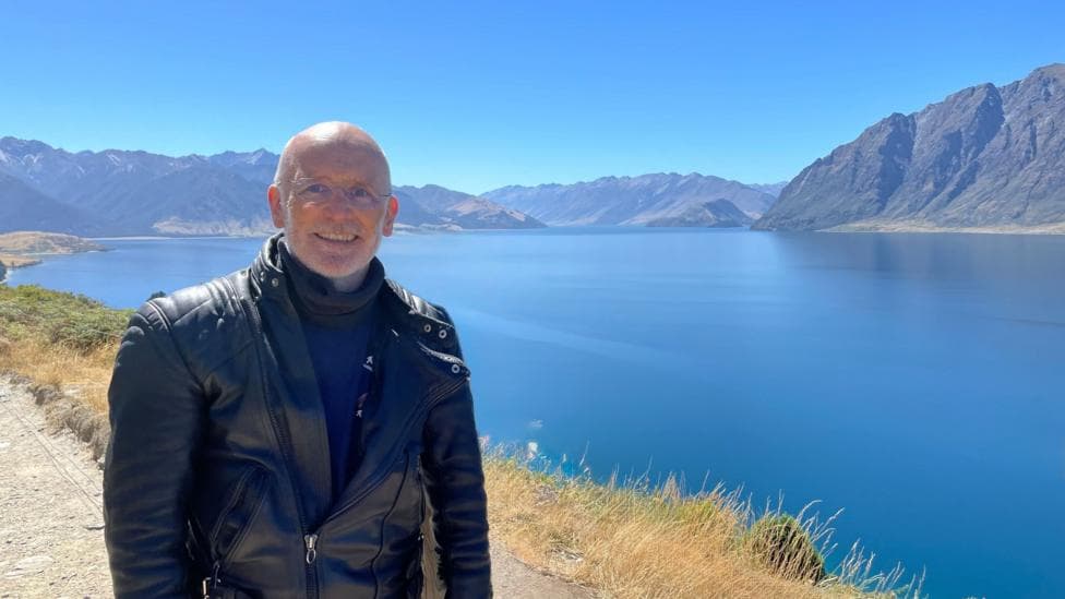 Sean Drysdale who is bald on top with white hair at the sides and a thin, light beard. He is wearing biker leathers and is standing in front a blue lake with mountains on the shore