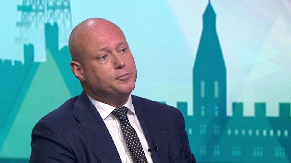 Dan Thomas being interviewed on Politics Wales . He is wearing a dark navy suit jacket, a white shirt and a dark tie with a light-coloured dot pattern. He has a bald head.