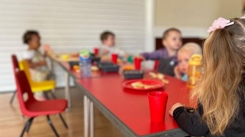 The image shows a group of unidentifiable toddlers sitting around a table eating and drinking. The table is red and the children's faces are blurry.