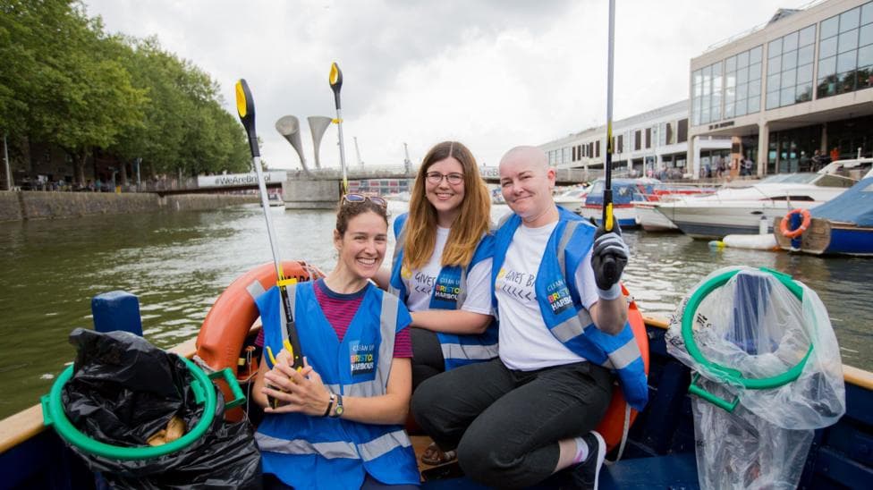 Three volunteers wearing blue high-visibility jackets sit on a boat sailing on Bristol Harbour as they hold up litter pickers to the camera. They are all smiling.