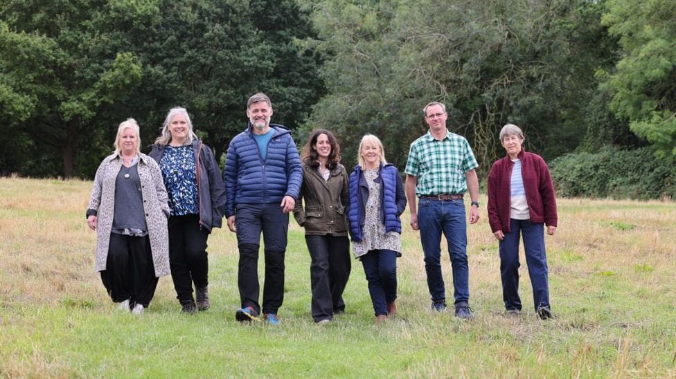 A group of seven men and women smile at the camera as they walk across a field. There are trees and bushes in the background.