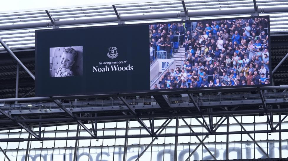 A big screen shows a picture of a smiling three-year-old boy, alongside the text: "In loving memory of Noah Woods." Alongside it on the screen football fans can be seen standing and applauding.
