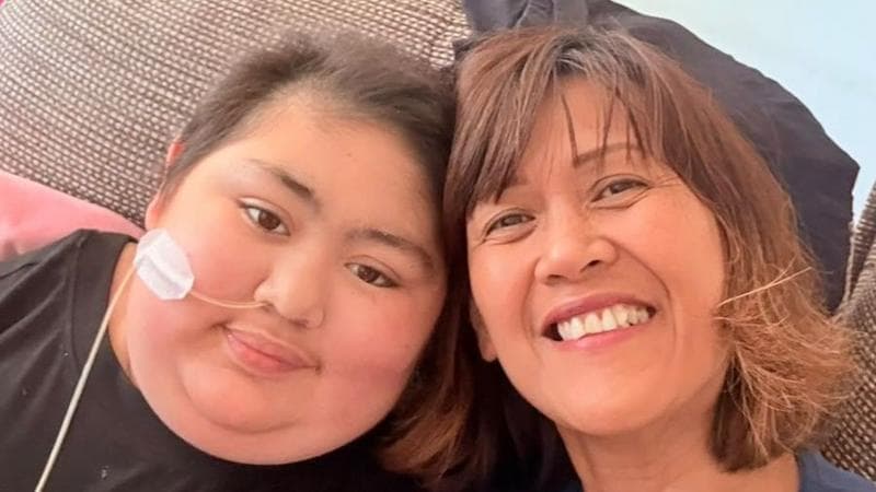 A woman and her daughter, sitting next to each other and smiling into a camera. The daughter, left, has short hair and has a tube inserted into her nose, which is taped to her face.