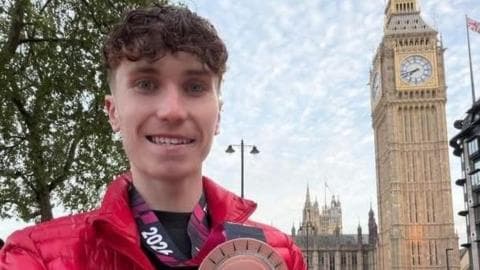 A man with brown hair and a red jacket, holding a medal in front of Big Ben