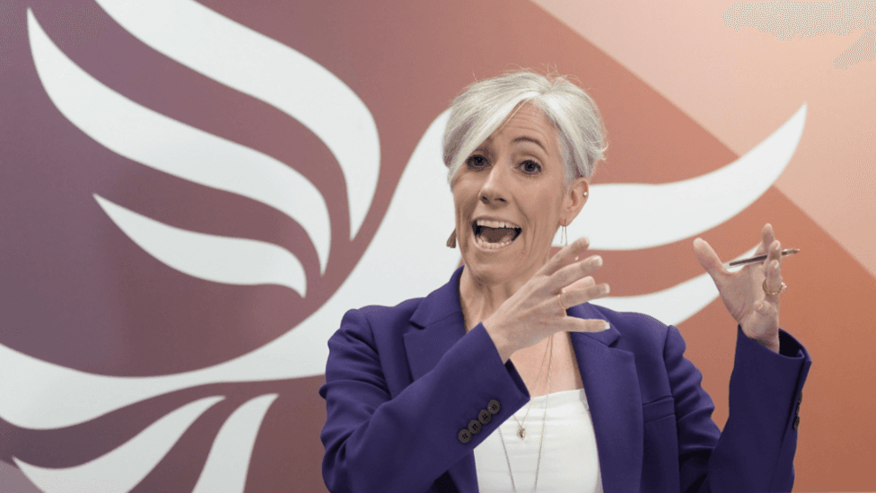 Daisy Cooper speaking on stage in front of a giant orange and white Liberal Democrat bird logo. She is gesticulating with her hands, wearing a purple jacket over a white top, and gold jewellery.