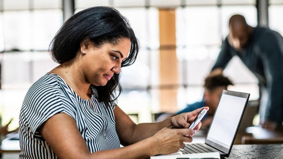 Woman looking at a phone sits at a desk with a laptop in front of her and a man and boy in the background.