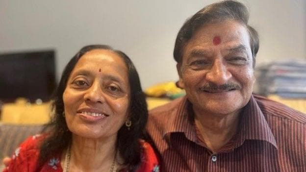 A hindu couple are looking straight at the camera and smiling. The woman is wearing a red top with flowers on it, she has thin black hair just past her shoulders and she has a small red dot called a bindi in the middle of her forehead. Her husband has his arm around here and is wearing a maroon verticle striped shirt and has a large bindi on his forehead.
