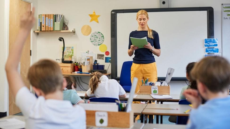 Stock image of a teacher teaching her class. She is wearing yellow trousers and a navy blue long-sleeved tshirt. Behind her is a Smart board and classroom items. In front of her are children learning at their desks - one boy has his hand up.