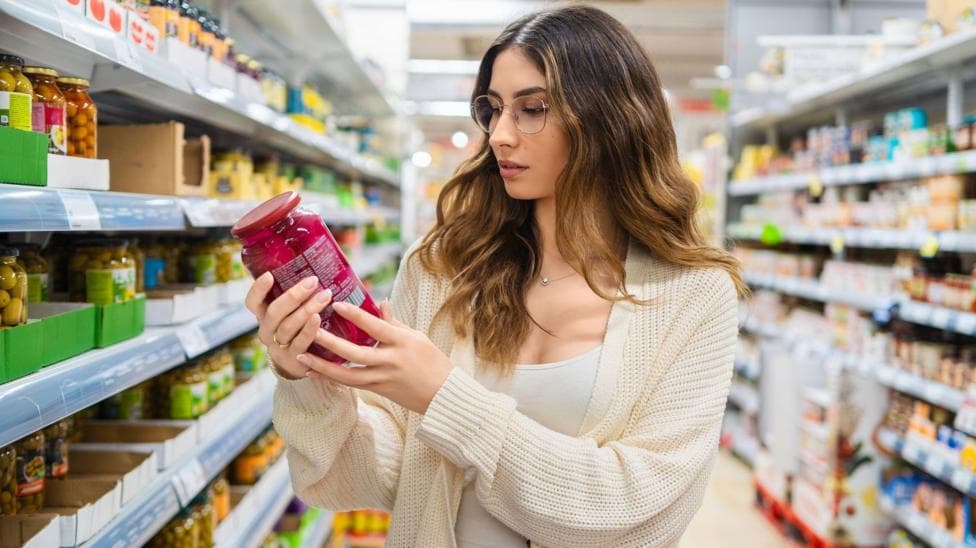 Young adult woman with glasses examining a jar in a grocery store aisle. She is focused on reading the label, surrounded by a variety of food products