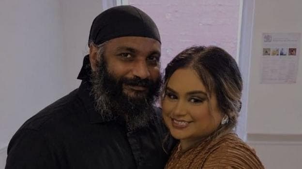 A mid-shot of a man and woman who are standing close together. The man has a long, black and grey beard and is wearing a black turban. The woman has dark brown hair with blonde highlights which is pulled back off her face. She has large diamante earrings and is wearing a bronze colour top. They ate both smiling.