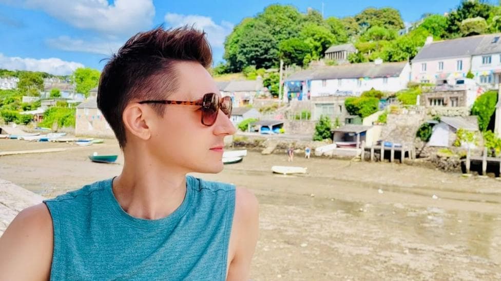 James Downs has short brown hair that is spiky on top. He is wearing a blue vest and brown sunglasses. He is looking out onto a river surrounded by boats and houses.