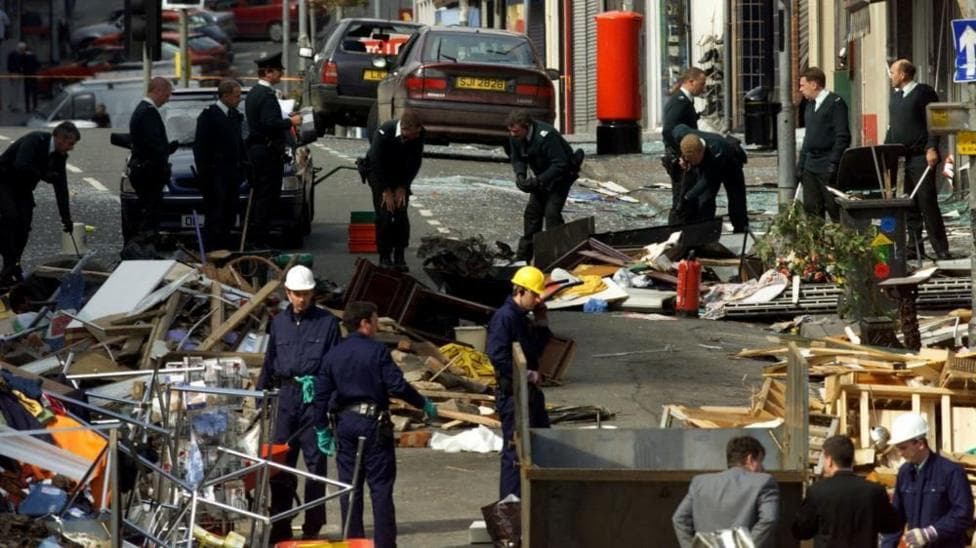 Security forces examine the scene of the Omagh bombing. There is rubble across the road and people wearing hard hats searching through.