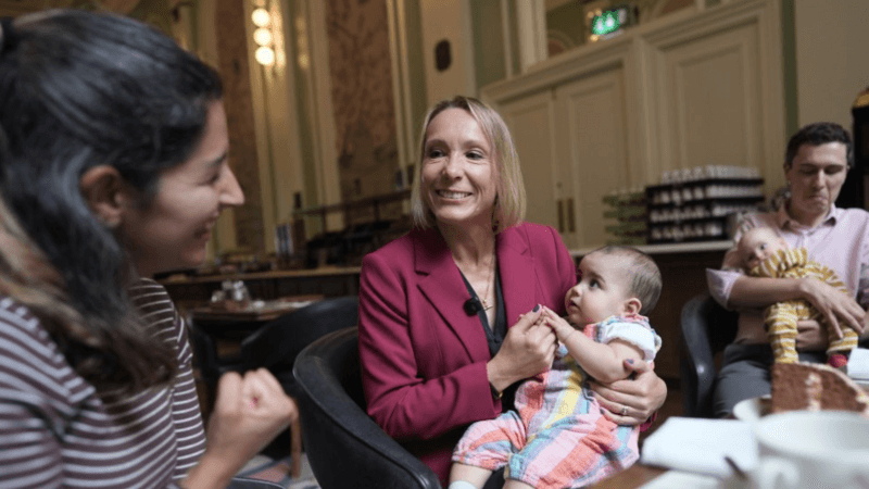 Helen Morgan is in the centre of the picture, smiling and holding a baby on her lap. Morgan has a blonde bob and is wearing a berry-coloured jacket over a navy top. She is looking at a woman in a stripy top on the left and a man holding another baby is on the right.