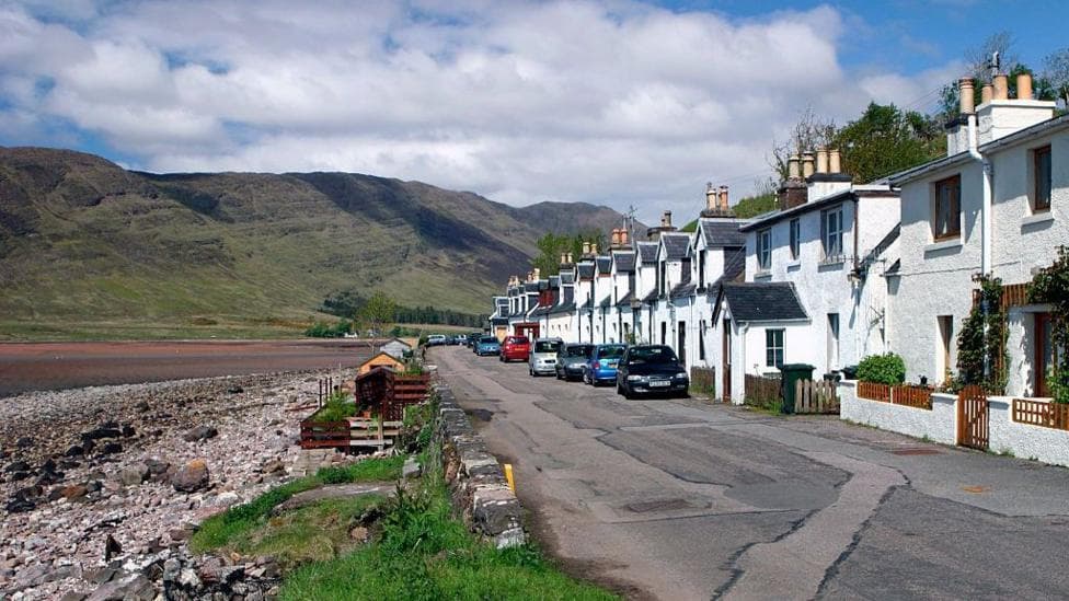 A row of white cottages is strung along a road that is right at the edge of a loch. There is a sandy bay with hills behind it in the distance.