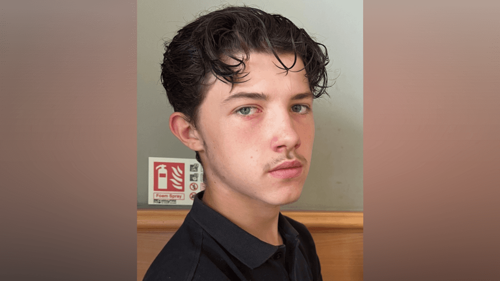 Declan Berry looks into the camera and has dark wavy hair and is wearing a black polo shirt