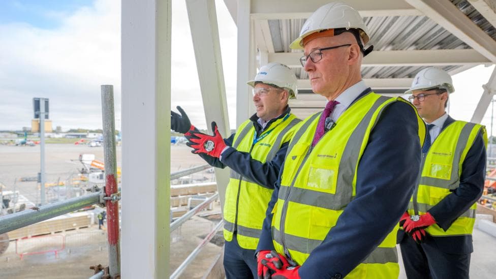 Three people wearing white hard hats, high-visibility yellow safety vests and protective gloves stand on an elevated construction platform overlooking an airport apron. Steel framework and scaffolding surround the group, while aircraft ground equipment and airport infrastructure are visible in the background.