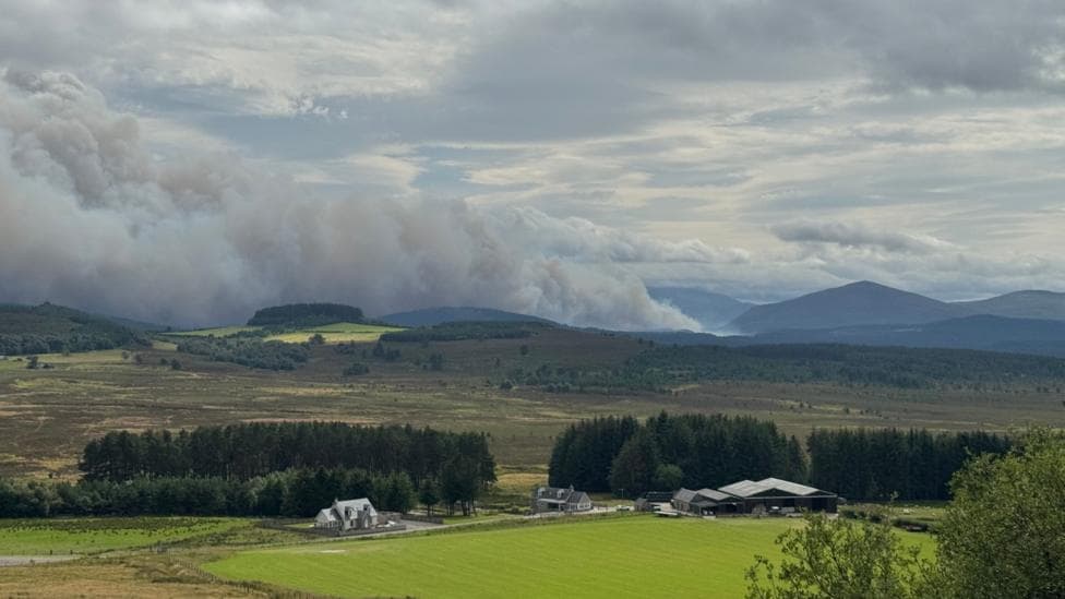 Large plume of smoke on the horizon. There are hills in the background, and trees, fields and houses in the foreground.