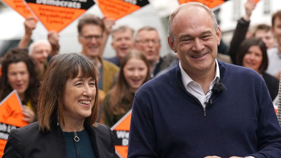 Jane Dodds is stood to the left of the picture and is shorter than Ed Davey, who is stood to the right. She has brown hair with a fringe, while he has short grey hair. They have a group of activists stood behind them all holding diamond shaped orange signs.