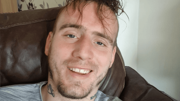 William Watson looking straight at the camera. He is sitting in an armchair. He is smiling with his teeth on show. He has long dark hair over his forehead and a short, dark beard. He has tatoos on either side of his neck and appears to be wearing a grey t-shirt.