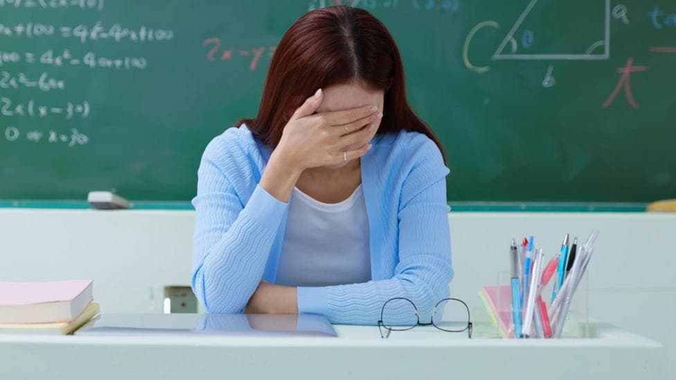 A woman with dark hair leans on a desk with her hand on her head, she is wearing a blue cardigan and white top