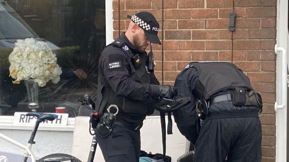 Two police officers in dark uniforms stand on a pavement outside a brick building, searching a dark bag. The officer in the foreground wears a cap with a black-and-white checked band, black gloves, body armour, a radio, and a body-worn camera, and looks down as he handles the bag with both hands. The second officer bends over beside him with his back to the camera, wearing a grey jacket and a utility belt. Behind them, a shop window holds a bouquet of white and pale-yellow hydrangeas, a red cup, and a white sign reading "RIP" followed by a partly hidden name. The edge of a bicycle with a blue saddle is visible in the bottom-left corner.