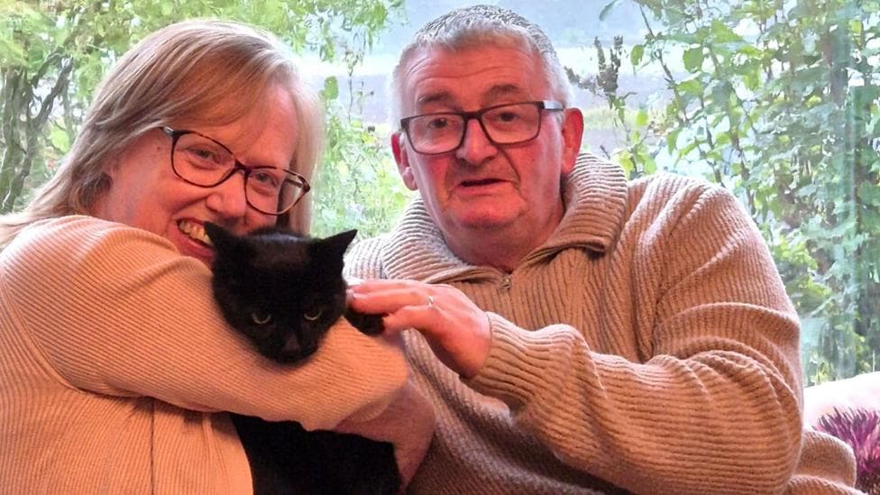 Mary and Billy Bowie, a man and woman, smiling at the camera and holding their black pet cat Pandora.