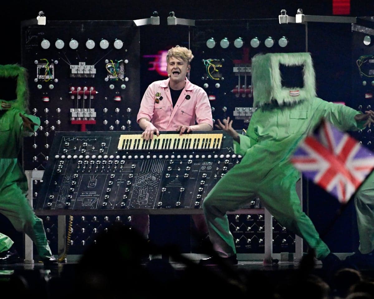 Sam Battle plays a synthesizer onstage between two dancers in furry green costumes at Eurovision