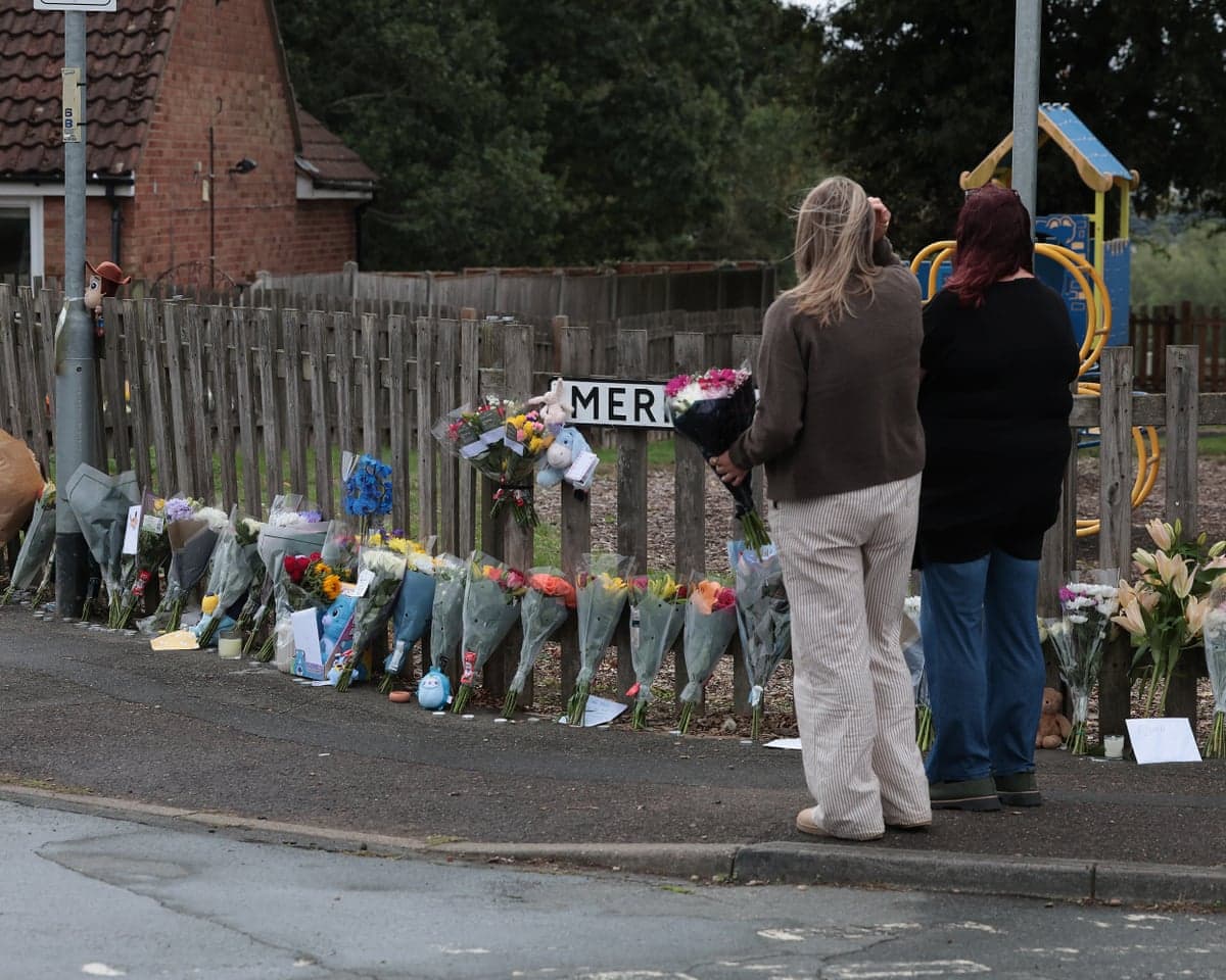 Two women, one holding a bunch of flowers, stand beside the playground and look at the flowers lining the fence