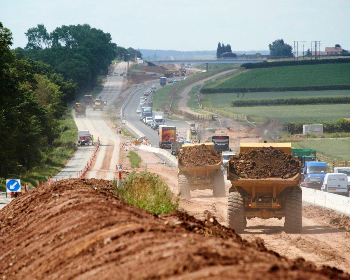 Construction vehicles move earth at road-widening site in England as traffic goes by