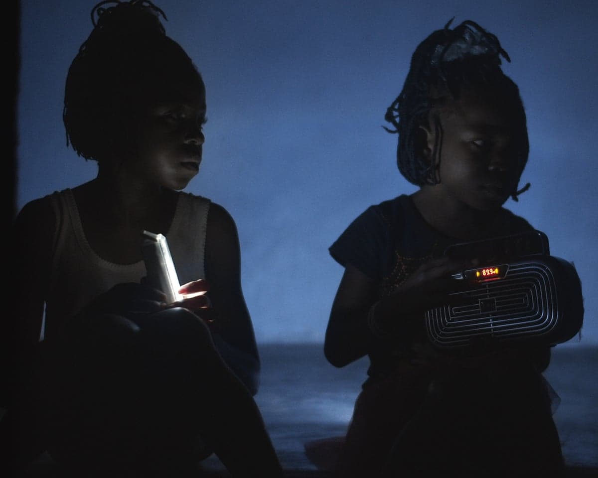 Two children sit in dim blue light, one holding a lit stick, the other holding a glowing radio in Rising Up at Night