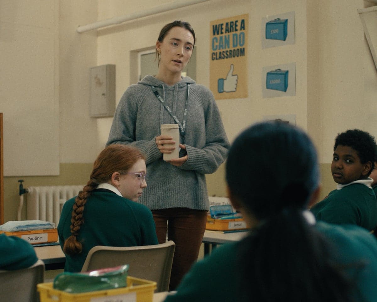 Film still from Bad Apples, showing a teacher, played by Saoirse Ronan, in a classroom wearing a grey hoodie and holding a drink in a plastic container.