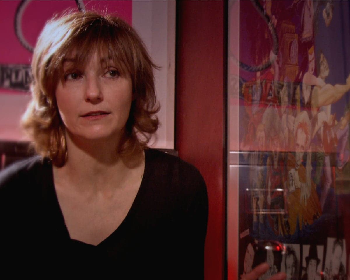 VivAlbertine with shoulder-length reddish hair in a black top stands near colorful punk-themed wall posters