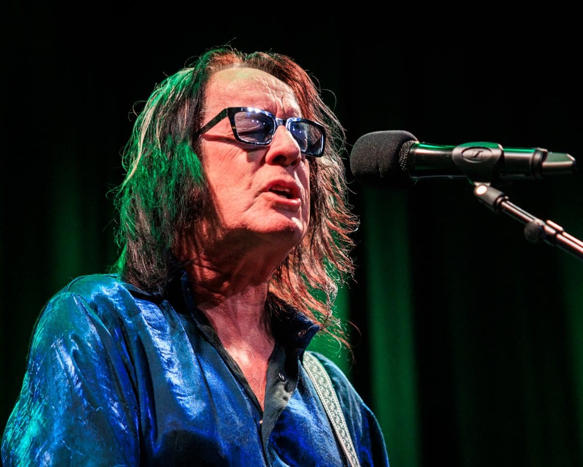 Todd Rundgren singing next to a microphone, wearing a blue bomber jacket