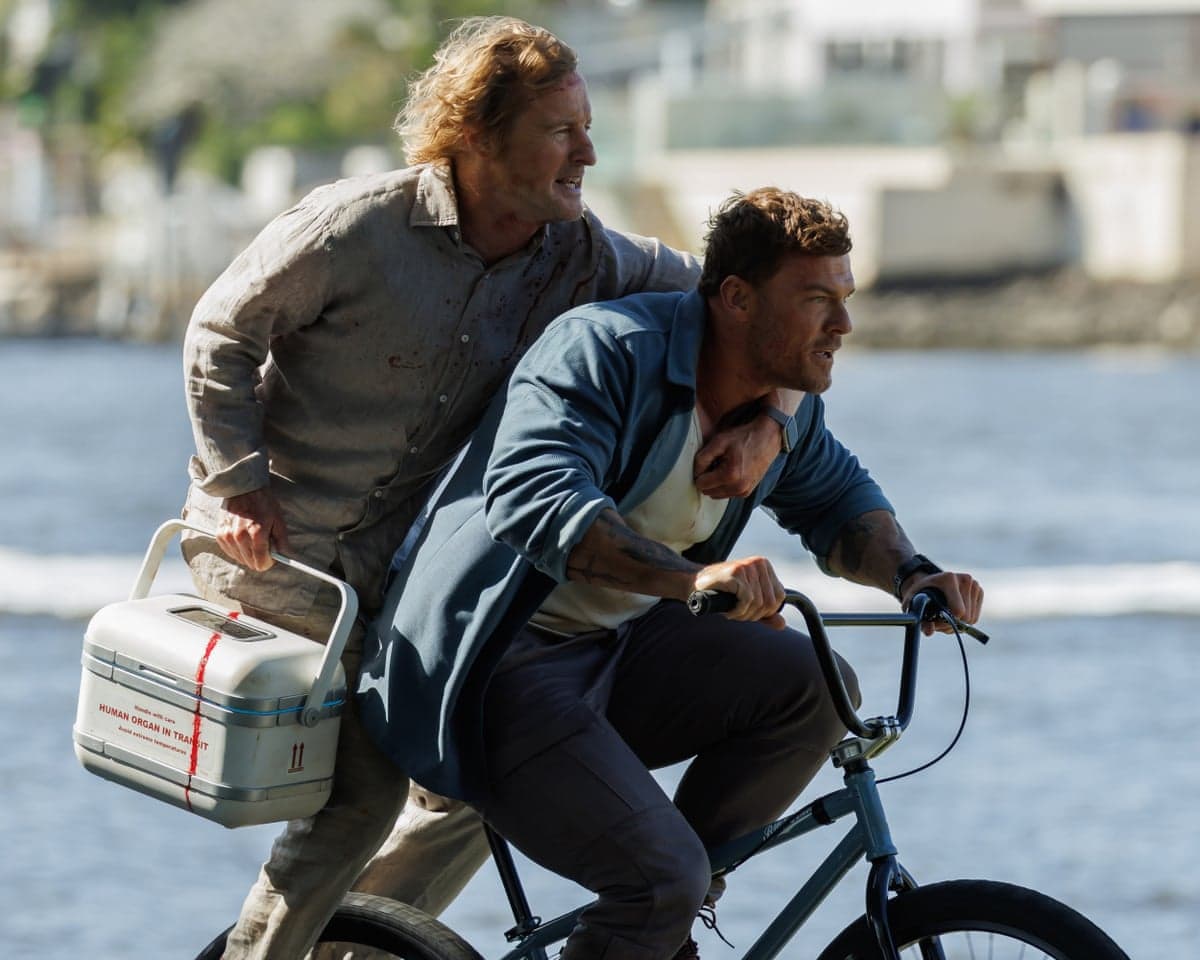 Owen Wilson and Alan Ritchsonride a bicycle, carrying a cooler labeled "human organ in transit"